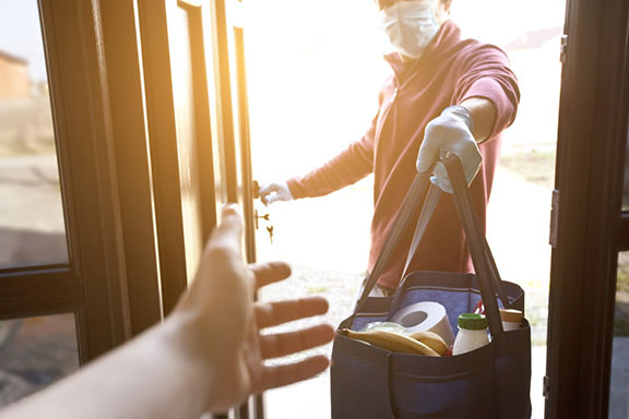 person wearing mask over mouth handing over grocery bag at house door