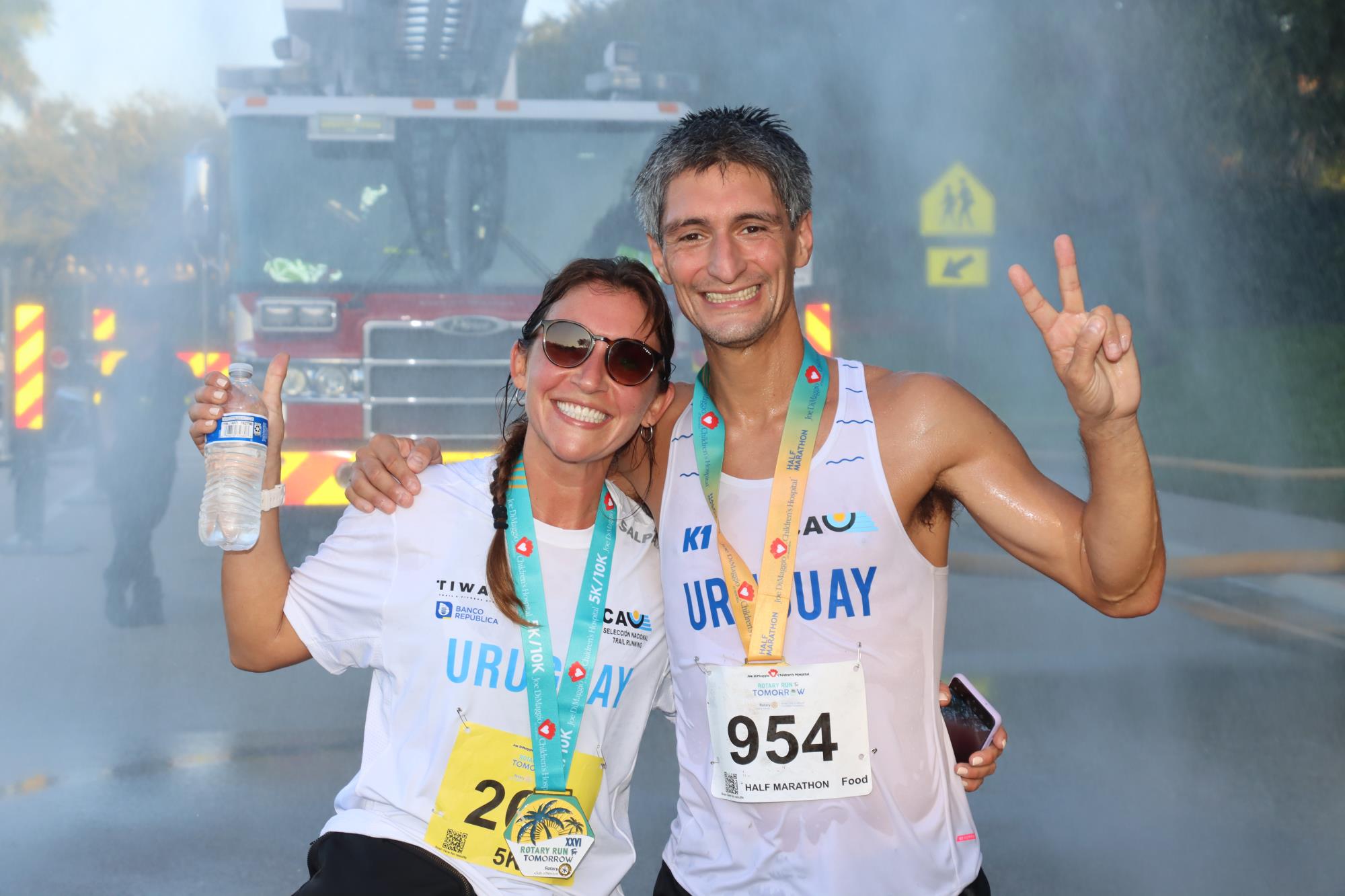 A woman and man wearing race bibs and medals from the 2023 Rotary Run races cool off under the Fire Engine hose shower after the race